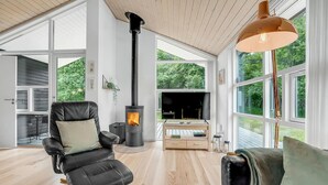 A modern living room with a black leather sofa, a wood-burning stove, and large windows offering a view of greenery. - Nice home in Asaa with kitchen (Asaa)