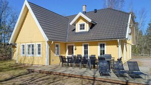 Une maison jaune avec un toit gris, une terrasse couverte avec des chaises et une cheminée.