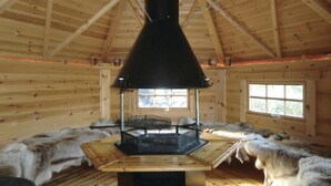 Interior de una cabaña de madera con un hogar central de leña, bancos cubiertos de pieles y una ventana que proporciona luz natural.