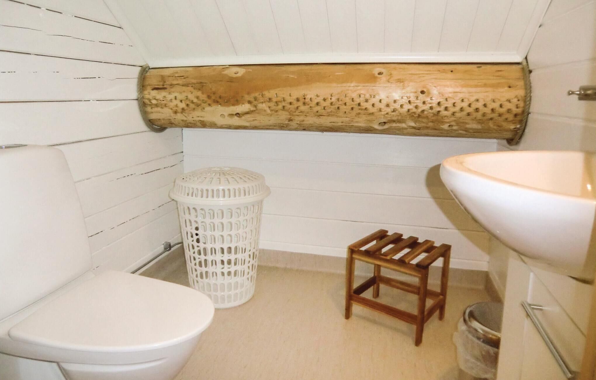 A small bathroom with a wooden log wall, a white toilet, a white trash bin, a wooden stool, and a white sink.
