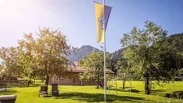 A flagpole with a flag, a building with a red-tiled roof, and a grassy area with trees and a mountain in the background.