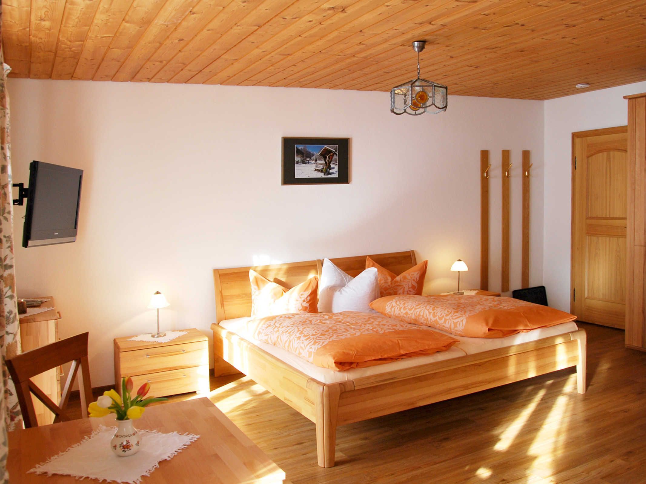 A bedroom with a wooden ceiling, a bed with orange bedding, a bedside table with a lamp, a picture on the wall, and a TV mounted on the wall.