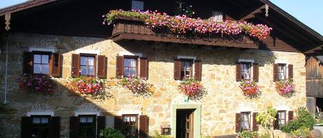 Un edificio de piedra con varias ventanas, algunas con contraventanas y balcones con flores.