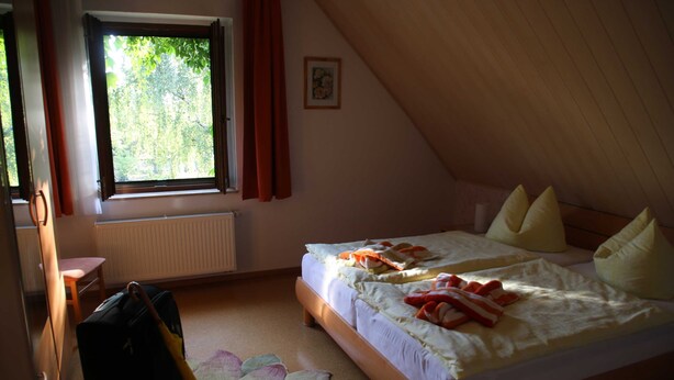 A bedroom with a bed, a window with curtains, a radiator, a chair, and a suitcase. - Apartment Blosenburgblick (Erfurt)