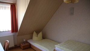 Quarto pequeno com cama de solteiro, teto inclinado, mesa de cabeceira com luminária e janela com cortinas.