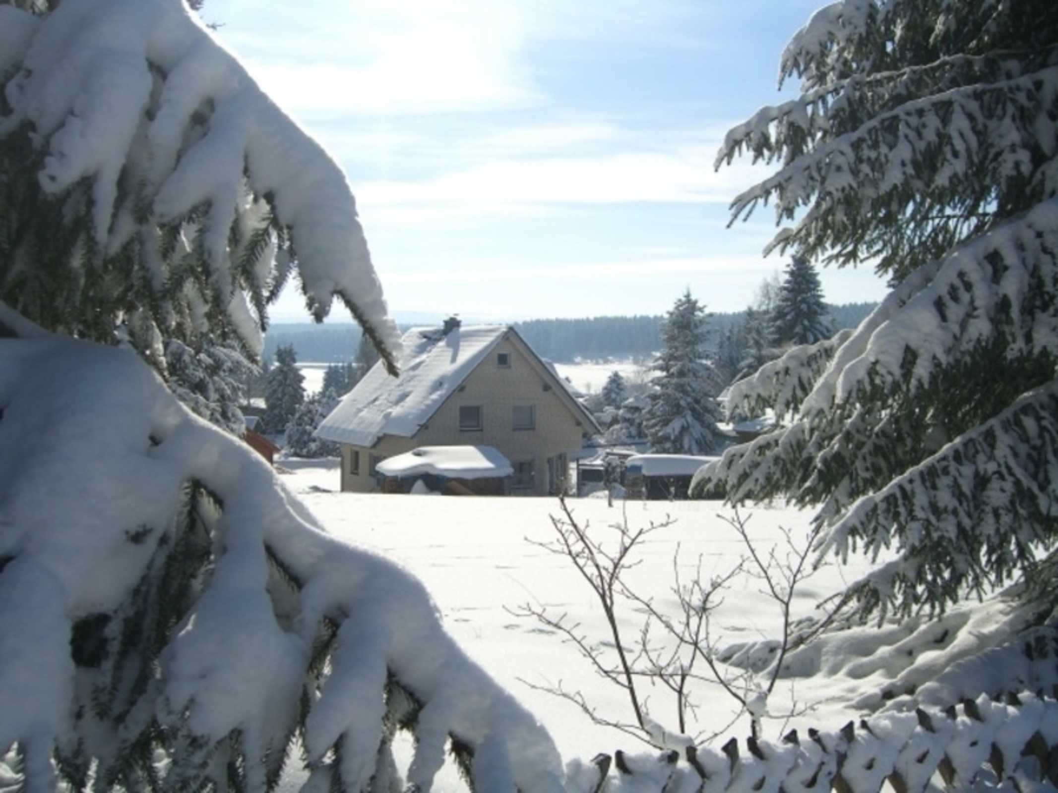 Une maison recouverte de neige, avec un toit en pente, entourée d’arbres chargés de neige et sous un ciel dégagé.