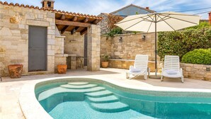 Un bâtiment en pierre avec une piscine, deux chaises longues blanches et un grand parasol.
