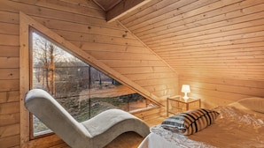 Acogedora habitación tipo cabaña de madera con techo inclinado, un ventanal grande, una butaca curvada, una mesita de noche con lámpara y una cama con almohadas.