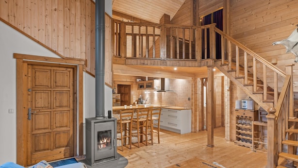 Un interior espacioso de madera con una chimenea central, una zona de cocina y una escalera que conduce a un nivel superior.