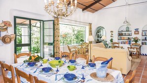 Un comedor con una mesa dispuesta para seis personas, rodeada de sillas de madera. Se observa una araña colgando sobre la mesa, y un estante lleno de libros al fondo.