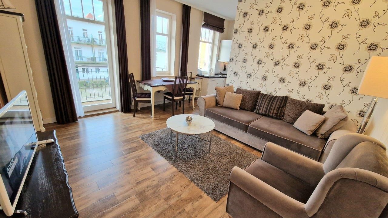 Ein Wohnzimmer mit Sofa, Sessel, Couchtisch, Fernseher, Esstisch, Stühlen und Blick ins Freie.