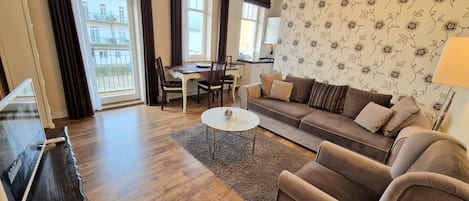 Ett vardagsrum med en soffa, fåtölj, soffbord, TV, matbord, stolar och utsikt över omgivningen.