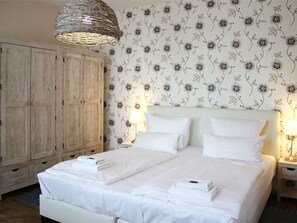 Un lit double avec du linge de lit blanc, une armoire en bois, un papier peint à motifs floraux et un luminaire suspendu.