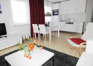 A modern living room with a white sofa, a coffee table with a vase of flowers, a dining area with white chairs, and a kitchen with white cabinets and appliances. - Villa Quisisana in the Kurstraße (EC) - Apartment Villa Quisisana (Graal-Müritz)