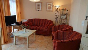 Una sala de estar con un sofá, un sillón y un televisor. Hay una mesa con un jarrón de flores.