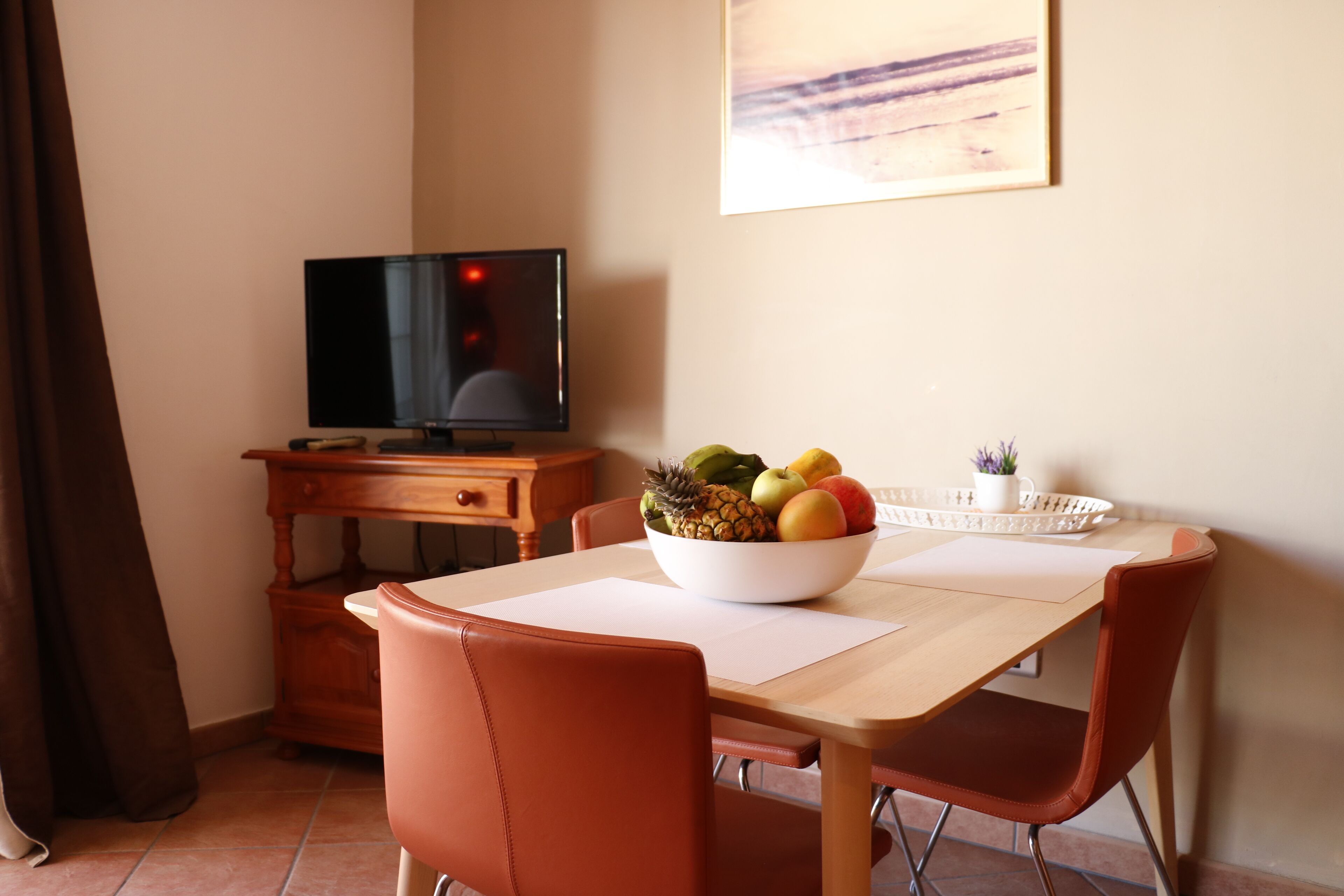 A dining area with a wooden table, two red chairs, a bowl of fruit, a television on a wooden stand, and a painting on the wall.