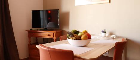 A dining area with a wooden table, two red chairs, a bowl of fruit, a television on a wooden stand, and a painting on the wall.