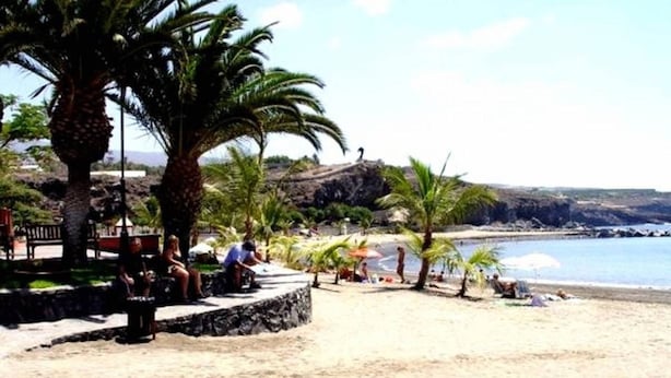 Una playa con palmeras, un promontorio rocoso y cielo despejado.