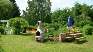 Un jardin agrémenté d’un barbecue en pierre, de bancs en bois et d’une pergola blanche.