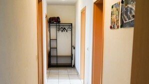 Un couloir étroit, avec un portant à vêtements sur le côté gauche et une valise posée à côté, et une porte ornée d’une affiche sur le côté droit.