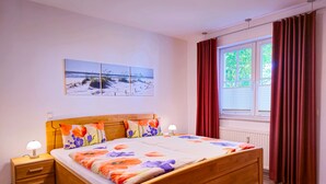 A bedroom with a wooden bed, two bedside tables, a nightstand, a window with curtains, and a beach scene painting on the wall. - 3 Room H 7 - Wohnpark Binz (with indoor pool) (Binz)