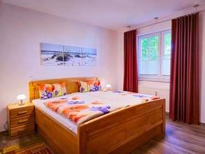 A bedroom with a wooden bed, two bedside tables, a nightstand, a window with curtains, and a beach scene painting on the wall. - 3 Room H 7 - Wohnpark Binz (with indoor pool) (Binz)