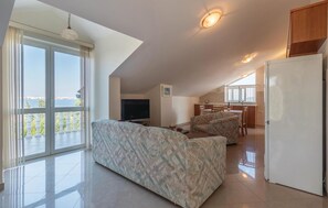 Ein großzügiger Wohnbereich mit einer großen Couch, einem Flachbildfernseher und einem Esstisch. Es gibt eine Küchenzeile mit Spüle und Kühlschrank. Das Zimmer verfügt über große Fenster mit Blick auf das Meer.