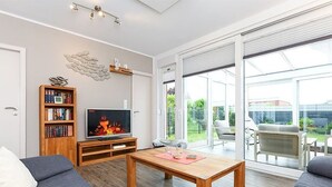A modern living room with a sofa, TV, wooden coffee table, bookshelf, and a sliding glass door leading to a balcony. - Holiday house Watt'n Huus (Dornum)