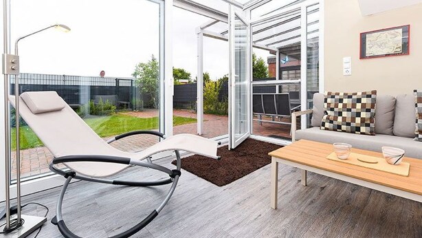 A modern living room with a glass door leading to a garden, a lounge chair, a sofa with patterned cushions, and a wooden coffee table. - Holiday house Watt'n Huus (Dornum)