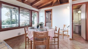 Uma sala de jantar com uma mesa posta para quatro pessoas, cadeiras de madeira e vista para o exterior através de grandes janelas.