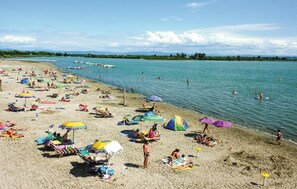 Una playa con numerosos paraguas y sillas de tomar el sol, un mar tranquilo y barcos a lo lejos.