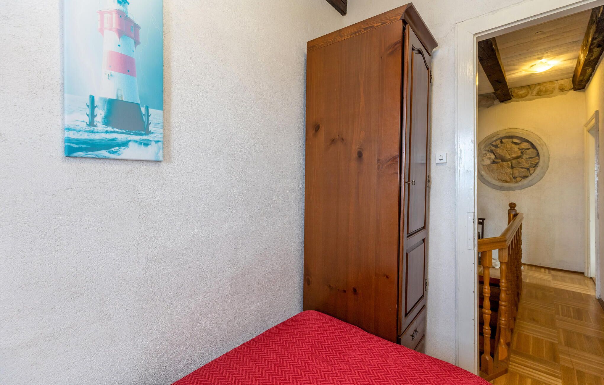 A room with a wooden wardrobe, a red bedspread, and a painting of a lighthouse.