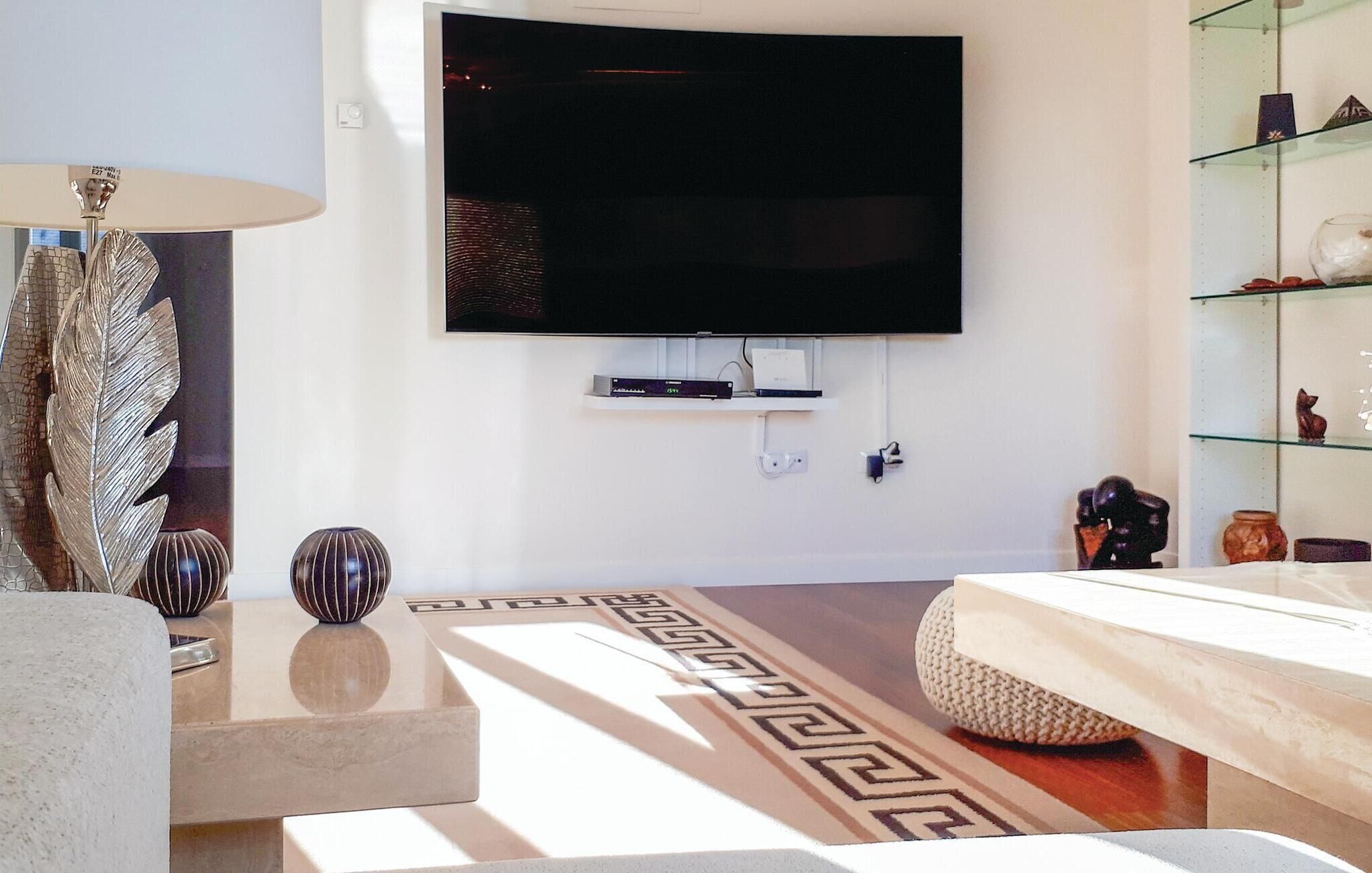 A modern living room with a flat-screen TV mounted on the wall, a decorative vase, a patterned rug, and a glass display shelf with various items.