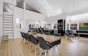 En rummelig spiseplads med et træbord og sorte stole, et køkken med moderne hårde hvidevarer, og et hyggeligt opholdsområde med en sofa og stole.