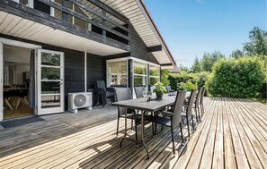 Et moderne hus med træterrasse, udendørs møbler og et stort vindue med udsigt over en have.