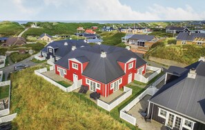 Ein rotes Haus mit schwarzem Dach, umgeben von weißen Zäunen, in einem Küstendorf mit weiteren Häusern und Blick auf das Meer.