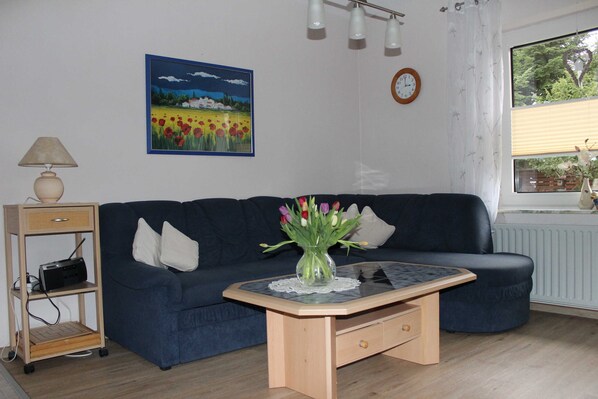 A living room with a dark blue sofa, a small wooden coffee table, a lamp, a picture frame, a clock, and a window with a view of trees.