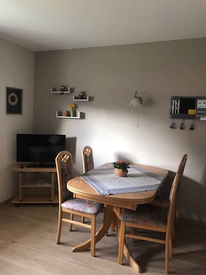Spiseområde med et rundt bord, stole, et fjernsyn og vægmonterede hylder med pynt.
