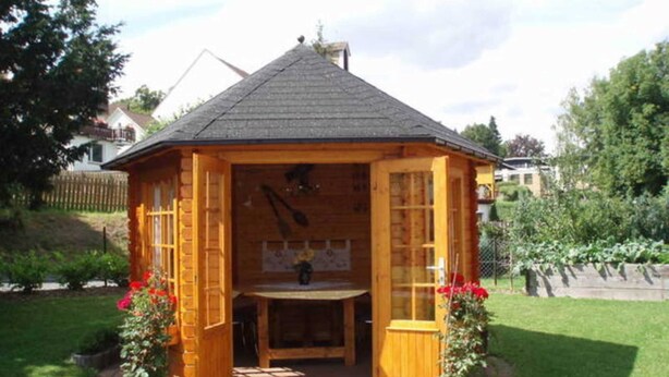A wooden gazebo with a tiled roof, a stone pathway leading to it, and potted plants on either side. - Vacation apartment (80 m²) - Gästehaus Müller (Walkenried)