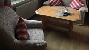 A cozy living room with a grey sofa, a wooden coffee table, and red and white striped cushions. A painting of flowers hangs on the wall, and a radiator is visible under the window. - Vacation apartment (80 m²) - Gästehaus Müller (Walkenried)