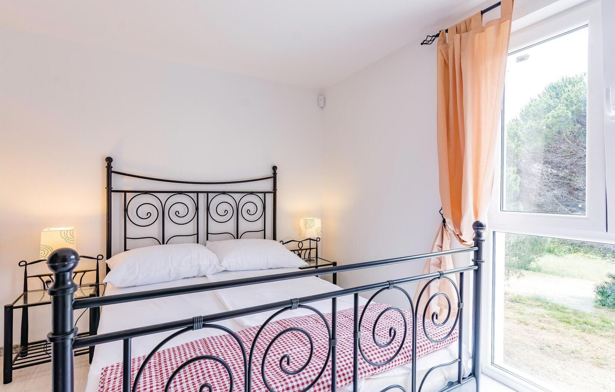 A bed with a metal frame, white bedding, and a checkered pillow. There are two bedside lamps and a window with curtains.