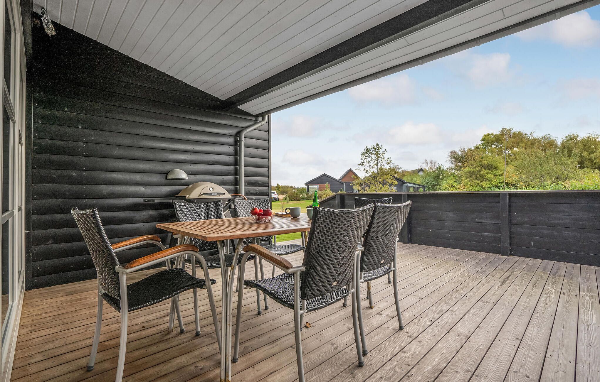 Una terraza de madera con una mesa y sillas, una lámpara y vistas a casas y follaje al fondo.