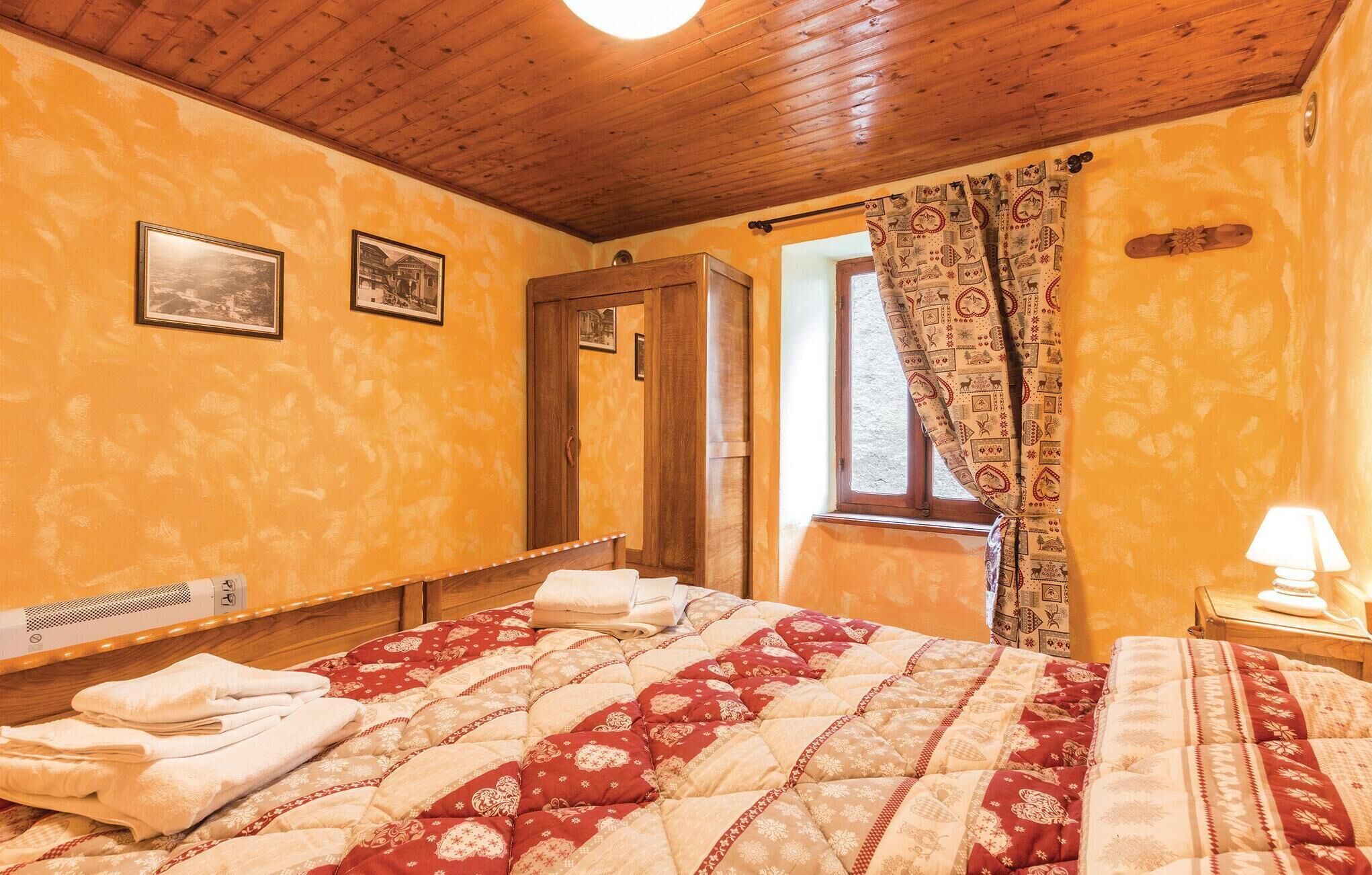 Ein Schlafzimmer mit Holzbalkendecke, einem Bett mit rotem und beigefarbenem Überwurf, einem Fenster mit gemusterten Vorhängen und einer Nachttischlampe.