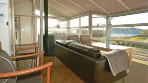 Ein gemütliches Zimmer mit Holzboden, einer Couch, zwei Sesseln, einem Tisch und einem großen Fenster mit Blick auf ein blaues Gebäude und Grünflächen.