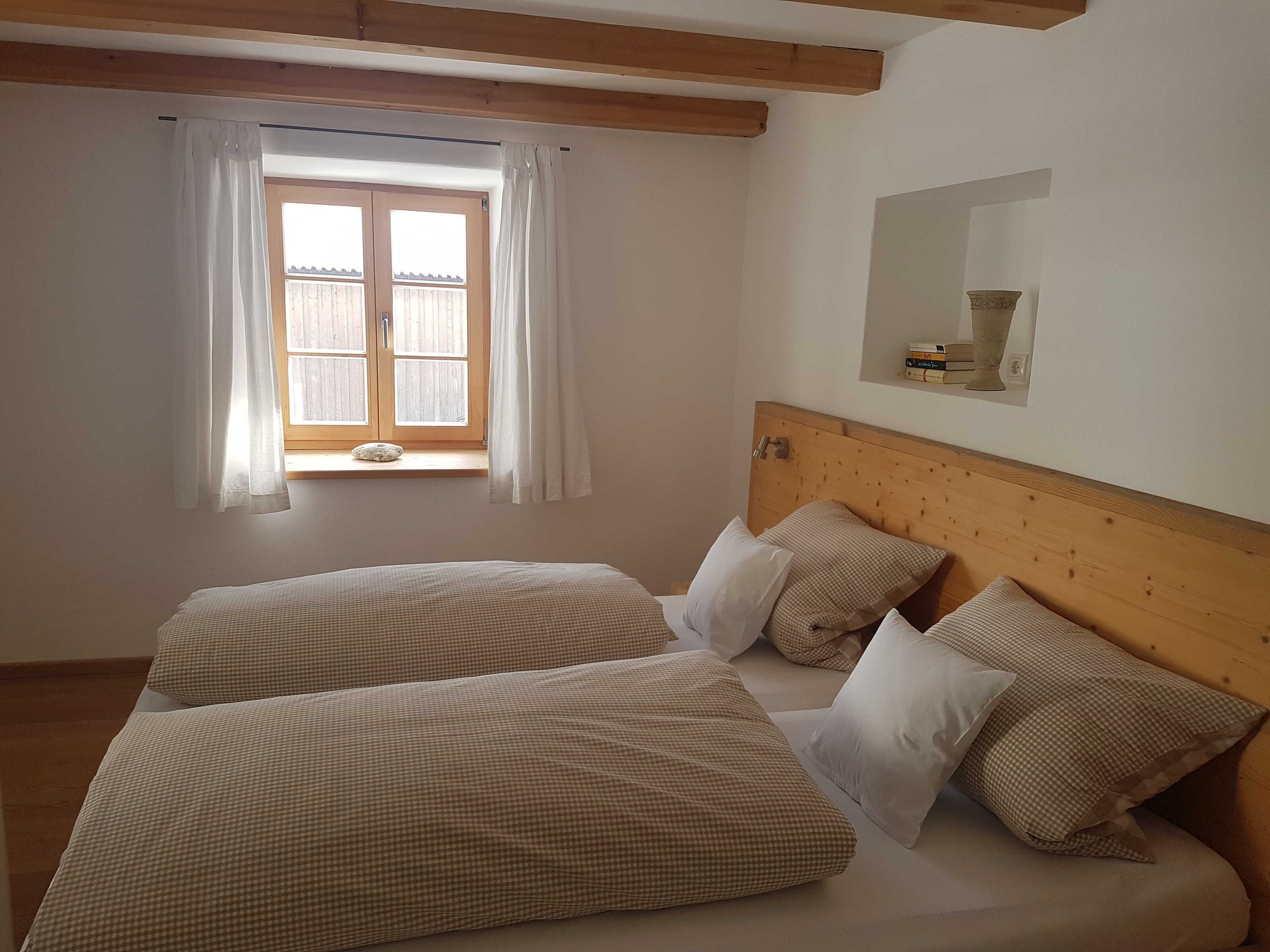 Ein Schlafzimmer mit zwei Betten, einem Fenster mit Vorhängen, einem Holz-Kopfteil und einem kleinen Regal mit Gegenständen.