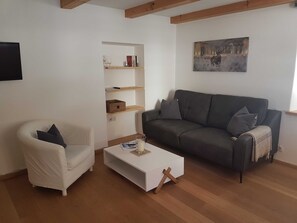 A living room with a white armchair, a grey sofa, a white coffee table, wooden shelves, and a painting on the wall. - Apartment G1 - Apartments Garhammer, Kreuth-glassworks (Kreuth)