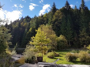 A wooden deck with a metal chair and a small table, surrounded by lush greenery and a forested backdrop. - Apartment G1 - Apartments Garhammer, Kreuth-glassworks (Kreuth)
