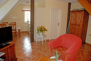 Una sala de estar con televisor, un sillón rojo, una mesita con un jarrón de flores y un comedor con sillas y una mesa.