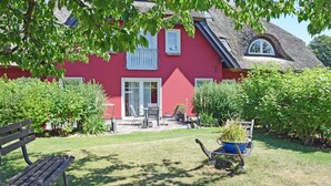 Une maison rouge avec un toit de chaume, un jardin avec un banc et une brouette remplie de plantes.