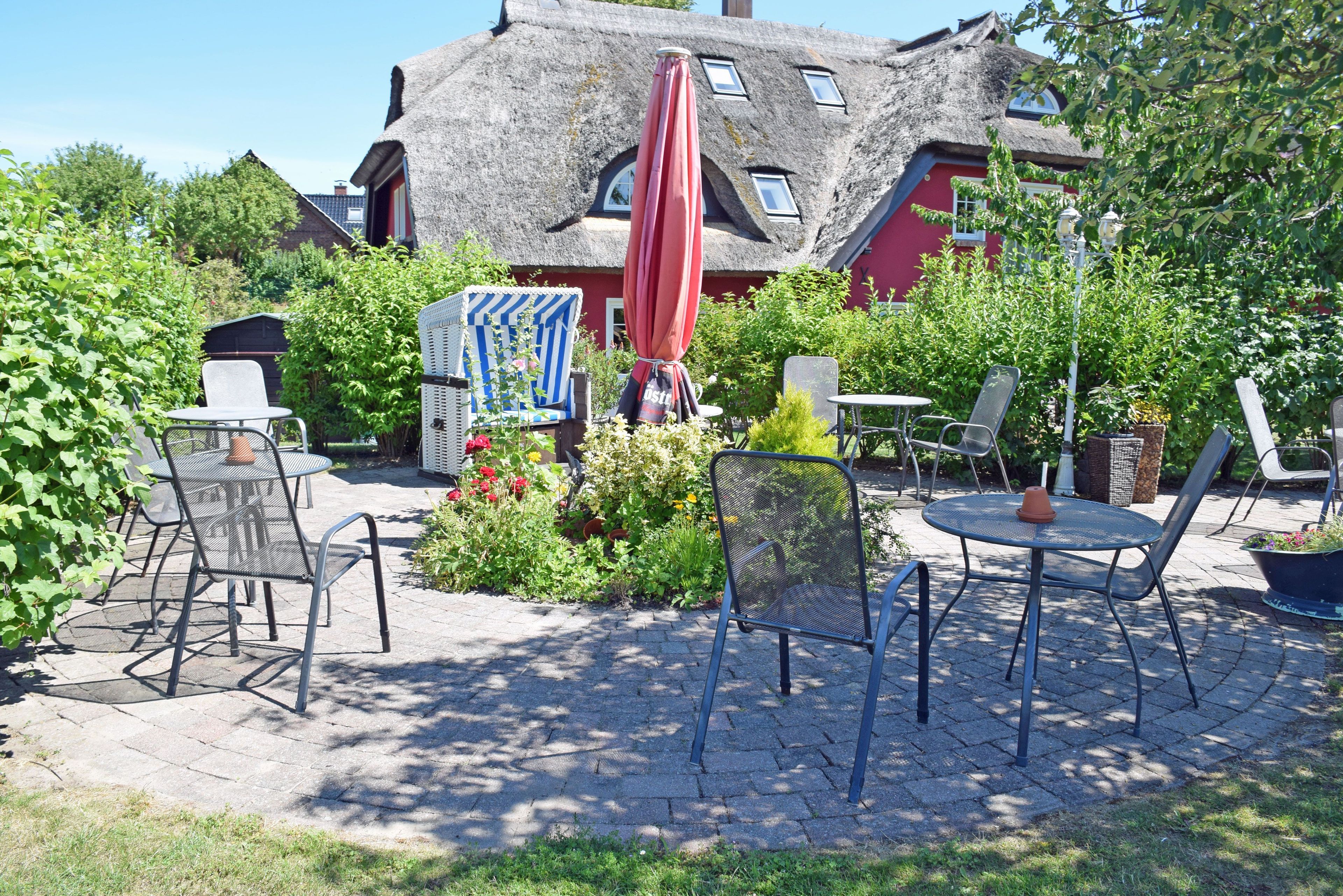 Een terras met tuinmeubilair, een rode parasol en een gebouw met rieten dak op de achtergrond.